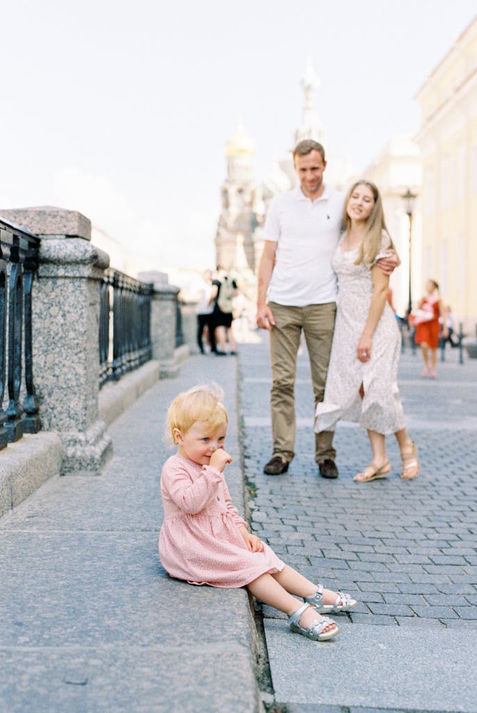 A toddler sits on a European street while parents stand nearby. Captures family moments.