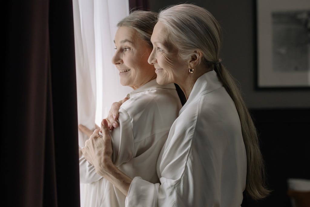 Two elderly women in white robes embracing near a window, symbolizing love and companionship.